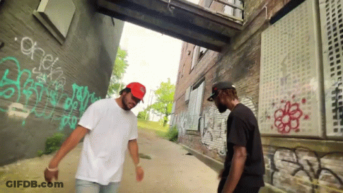 Two Men Are Standing In Front Of A Brick Wall With Graffiti On It Including One That Says Check GIF