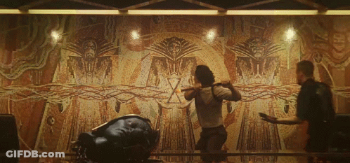 Two Men Are Standing In Front Of A Large Wall With A Drawing Of A Man With A Sword GIF