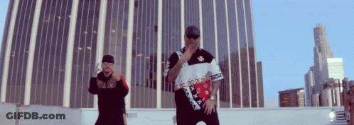 Two Men Are Standing In Front Of A Tall Building And One Has A Shirt That Says ' Tommy Hilfiger ' On It GIF