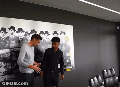Two Men Are Standing In Front Of A Wall With A Picture Of A Group Of Men GIF
