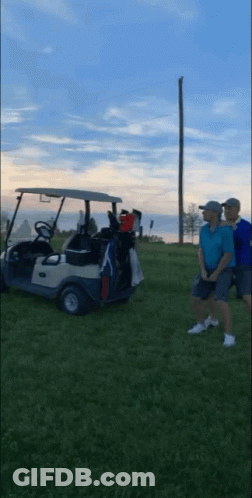 Two Men Are Standing Next To A Golf Cart On A Grassy Field GIF