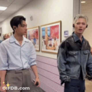 Two Men Are Standing Next To Each Other In A Hallway With Paintings On The Wall . GIF