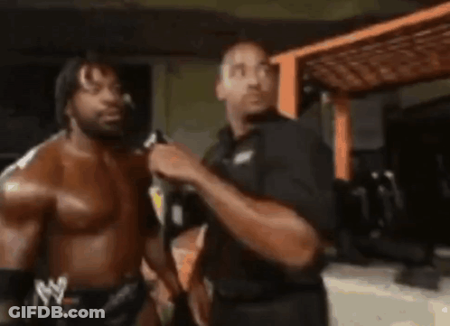 Two Men Are Standing Next To Each Other In A Room . One Of The Men Is Wearing A Shirtless Wrestling Outfit . GIF