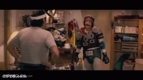 Two Men Are Standing Next To Each Other In A Room . One Of The Men Is Wearing A Superhero Costume . GIF