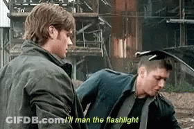 Two Men Are Standing Next To Each Other In Front Of A Building . One Of The Men Is Holding A Flashlight . GIF