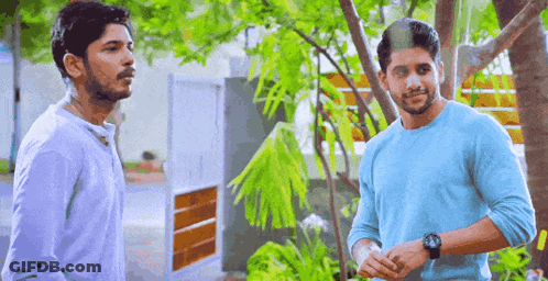 Two Men Are Standing Next To Each Other In Front Of A Tree Talking To Each Other . GIF