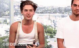 Two Men Are Standing Next To Each Other In Front Of A Window . One Of The Men Is Wearing Suspenders And A Tank Top . GIF