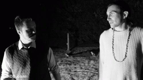 Two Men Are Standing Next To Each Other In The Dark And One Has A Necklace Around His Neck GIF