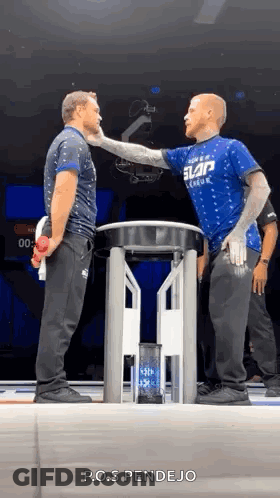 Two Men Are Standing Next To Each Other On A Stage And One Of Them Is Wearing A Blue Shirt That Says Slap League . GIF