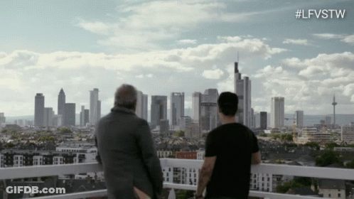 Two Men Are Standing On A Balcony Looking At A City Skyline With The Hashtag #Lfvstw GIF