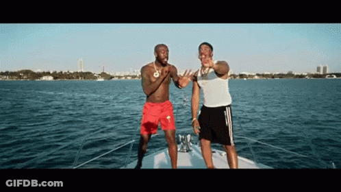 Two Men Are Standing On A Boat In The Water . One Of The Men Is Wearing Red Shorts . GIF