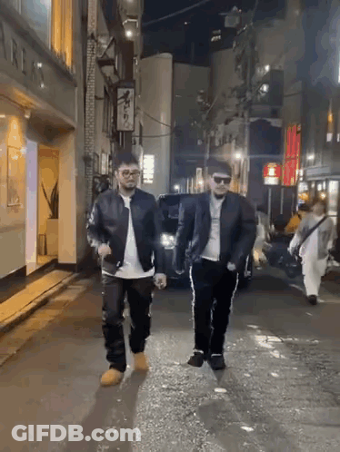 Two Men Are Walking Down A City Street At Night GIF