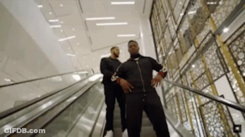 Two Men Are Walking Down An Escalator In A Building . GIF