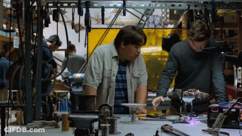 Two Men Are Working On A Project In A Workshop And One Of Them Is Looking At A Box That Says Welding GIF