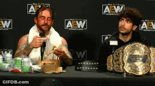 Two Men At Table In Front Of AEW Wrestling Wall GIF
