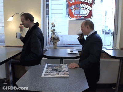 Two Men At Table In Front Of Bakery Sign GIF