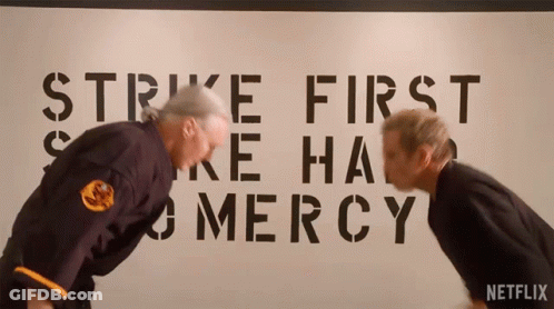 Two Men Bending Over In Front Of Strike First And Mercy Sign GIF