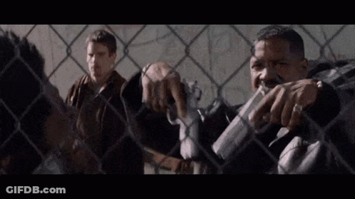 Two Men By Chain Link Fence, One With Gun GIF