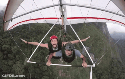 Two Men Flying Hang Glider Over Green Hillside GIF