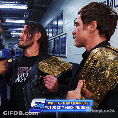 Two Men Holding Wrestling Belts In Front Of A WWE Tag Team Champions Motor City Machine Guns Banner GIF