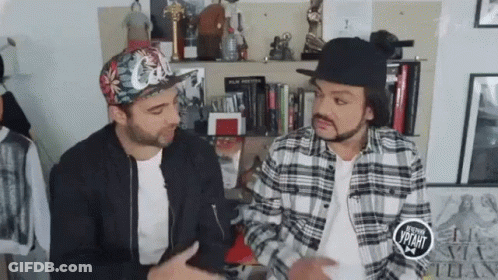 Two Men In Hats And Plaid Shirts Sitting By Bookshelf GIF