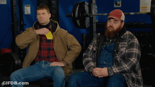 Two Men In Plaid Shirts And Overalls Are Sitting In A Gym One Is Holding A Yellow Card GIF