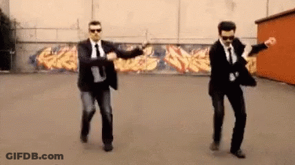 Two Men In Suits And Ties Are Dancing In A Parking Lot In Front Of A Graffiti Wall . GIF