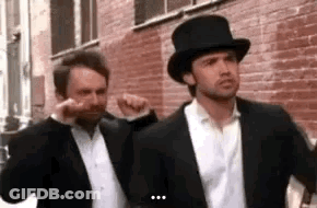 Two Men In Suits And Top Hats Are Standing Next To Each Other On A Sidewalk . GIF