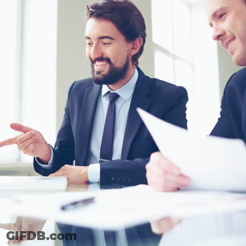 Two Men In Suits Are Sitting At A Table Looking At A Piece Of Paper GIF