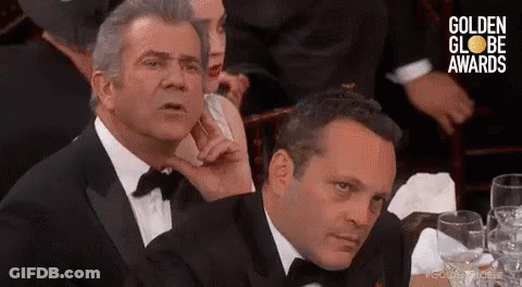 Two Men In Tuxedos Are Sitting At A Table With Glasses Of Wine And Looking At Something . GIF