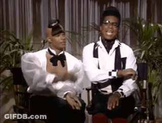 Two Men In Tuxedos Sitting In Director's Chairs Talking GIF