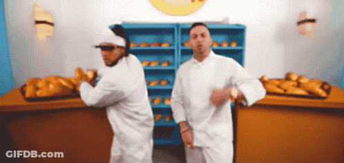 Two Men In White Chefs Uniforms Are Dancing In Front Of A Bunch Of Bread GIF