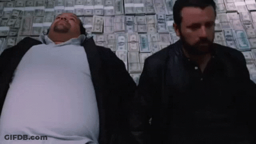 Two Men Laying On Top Of A Pile Of Money With One Of Them Having A Beard GIF