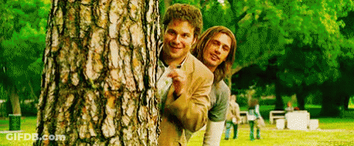 Two Men Peeking Behind Tree In Park GIF