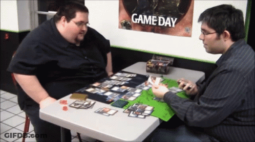 Two Men Playing A Game Of Cards With A Game Day Poster Behind Them GIF