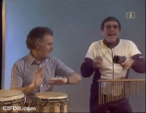 Two Men Playing Drums In Front Of BBC TV Screen GIF