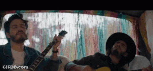 Two Men Playing Guitars In Van With Colorful Curtain GIF