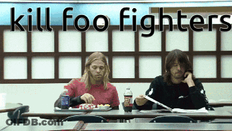 Two Men Sit At A Table In Front Of A Wall That Says Kill Foo Fighters On It GIF