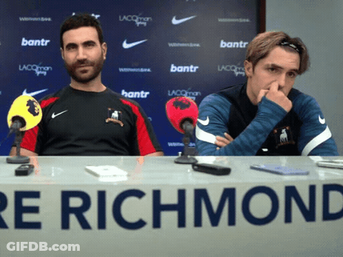 Two Men Sit At A Table With A Sign That Says Richmond GIF