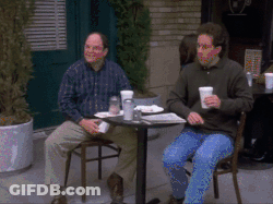 Two Men Sit At A Table With Cups In Their Hands GIF