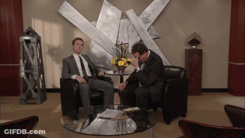 Two Men Sit In Chairs In Front Of A Table With A Vase Of Flowers GIF