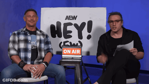 Two Men Sit In Front Of A Sign That Says " Hey " GIF