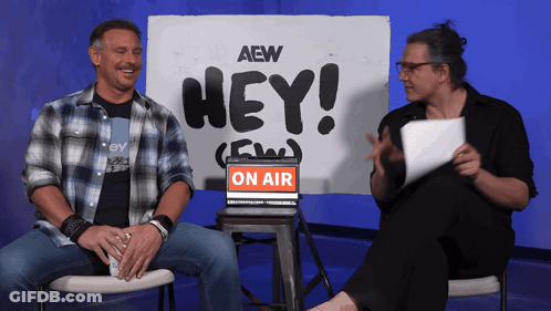 Two Men Sit In Front Of A Sign That Says Hey On Air GIF