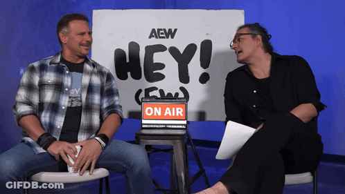 Two Men Sit In Front Of A Sign That Says Hey On Air GIF
