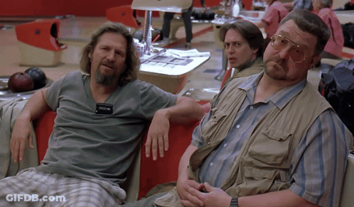 Two Men Sit On A Couch In A Bowling Alley With A Bowling Ball Behind Them GIF