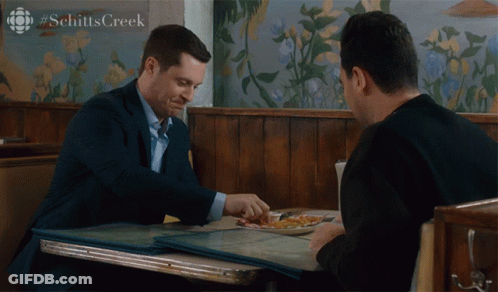 Two Men Sitting At Schitt's Creek Sign GIF