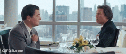 Two Men Sitting At Table In Front Of Large Window Talking GIF