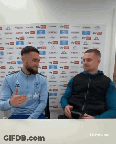 Two Men Sitting In Front Of Sky Bet Wall GIF