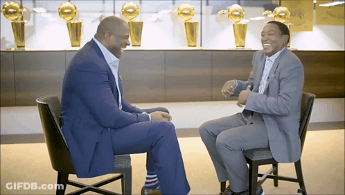 Two Men Sitting In Front Of Trophy Wall GIF
