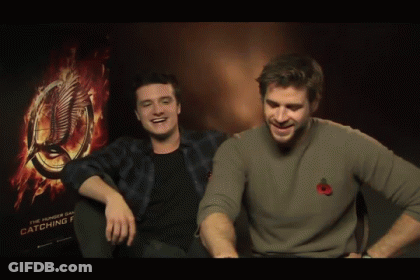 Two Men Sitting Next To Each Other In Front Of A Hunger Games Poster GIF